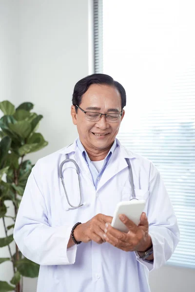 Handsome old doctor in white medical coat and eyeglasses is using a smartphone and smiling while standing against window