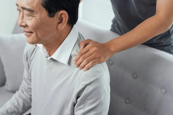 Asian old man shoulder pain, sitting on sofa , son massaging father shoulder.