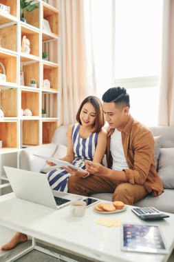 Gülümseyen genç çift, faturaları ve ev bakımı dizüstü bilgisayar kullanarak kanepede oturup, evrak okumak