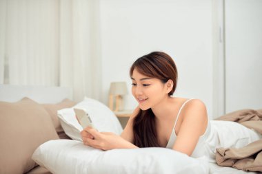 Genç Asyalı kadın yatağında cep telefonu kullanıyor. Yüzü temiz ve yatakta uzanıyor..