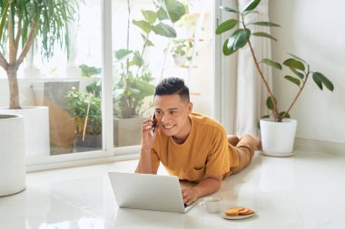 Gülümseyen adam cep telefonuyla konuşuyor ve evde yerde yatarken dizüstü bilgisayar kullanıyor.