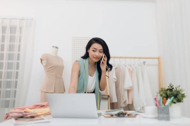 Genç Asyalı moda tasarımcısı / iş adamı laptopunu kullanıyor ve stüdyosunu arıyor.