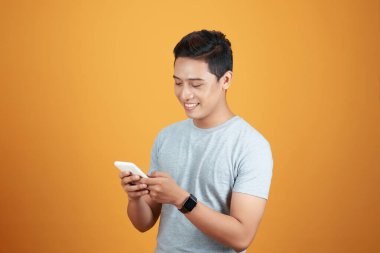 Akıllı telefon kullanan yakışıklı Asyalı genç adam, stüdyonun turuncu arka planında vuruldu.