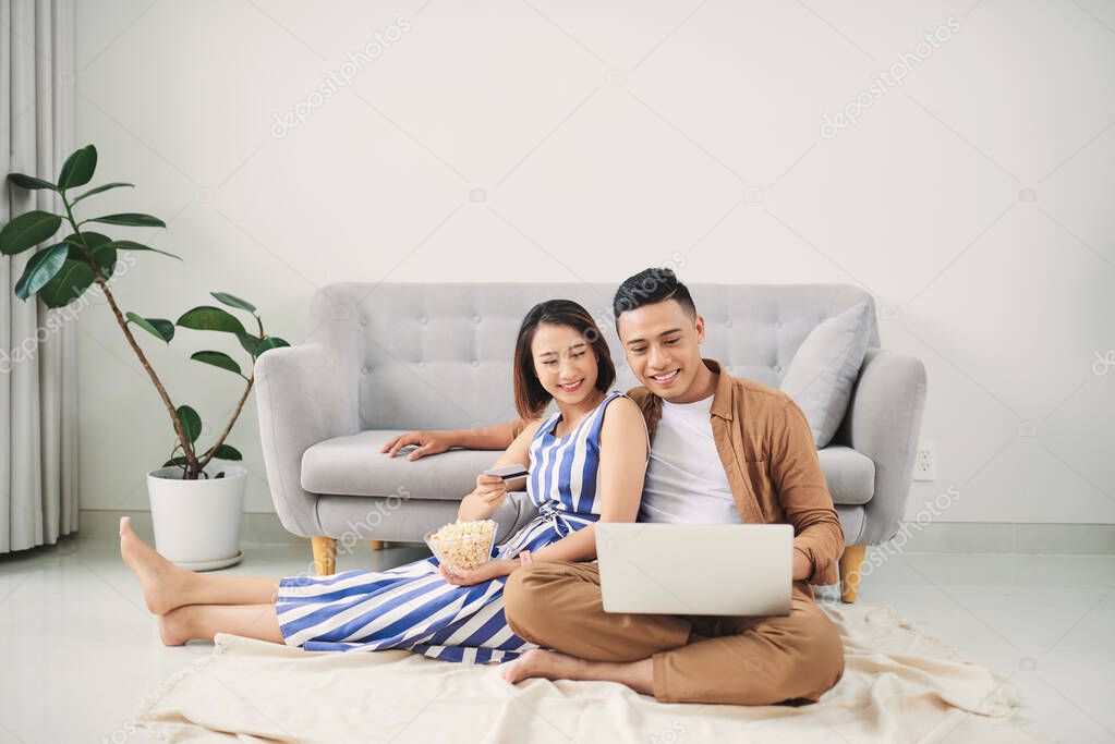 Beautiful young woman holding credit card and looking at laptop with smile while sitting together with young man