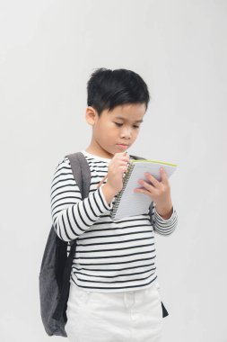 Genç Asyalı okul çocuğu beyaz arka planda sırt çantasıyla not defterine yazıyor.