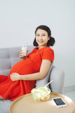 Kanepede otururken salata yiyip süt içen güzel, genç Asyalı hamile kadın..