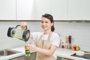 Keten önlüklü genç bir kadın sebze ve yeşilliklerle çevrili blenderdan şişeye yeşil smoothie dolduruyor..