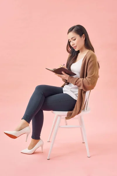 Happy young woman is sitting in a chair thinking about something, writing everything in a notebook