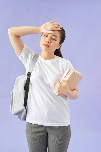 Tired young woman student with backpack on purple background.  Hold books keeping eyes closed put hand on head