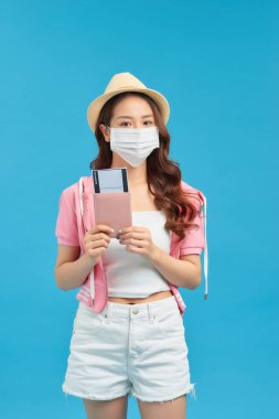 Pasaportlu ve mavi arka planda bileti olan el yapımı pamuk maskeli genç bir gezgin kadın. Coronavirus sırasında trip konsepti.