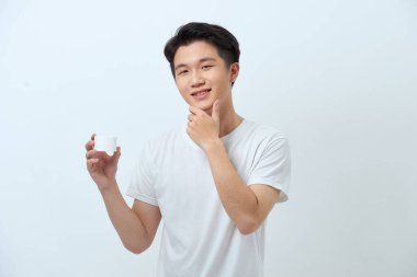 Genç adam çenesine dokunuyor, elinde yüz kremiyle kameraya gülümsüyor.