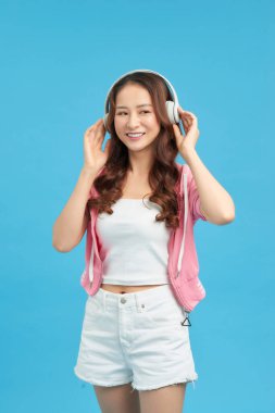 Sevimli Asyalı kadın şarkı söyleyip eğlenirken mavi arka planda izole edilmiş kablosuz kulaklıklar kullanıyor.