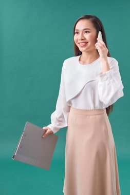 Elinde klasör olan genç bir kadın, turkuaz arka planda cep telefonuyla konuşuyor.