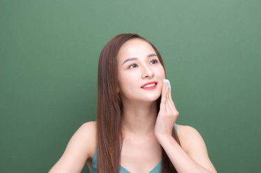 Yeşil arka planda izole edilmiş pamukla yüzünü temizleyen gülümseyen Asyalı genç kadının portresi..