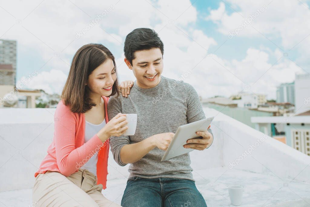 Young Asian couple using tablet and drinking tea/coffee together on rooftop.