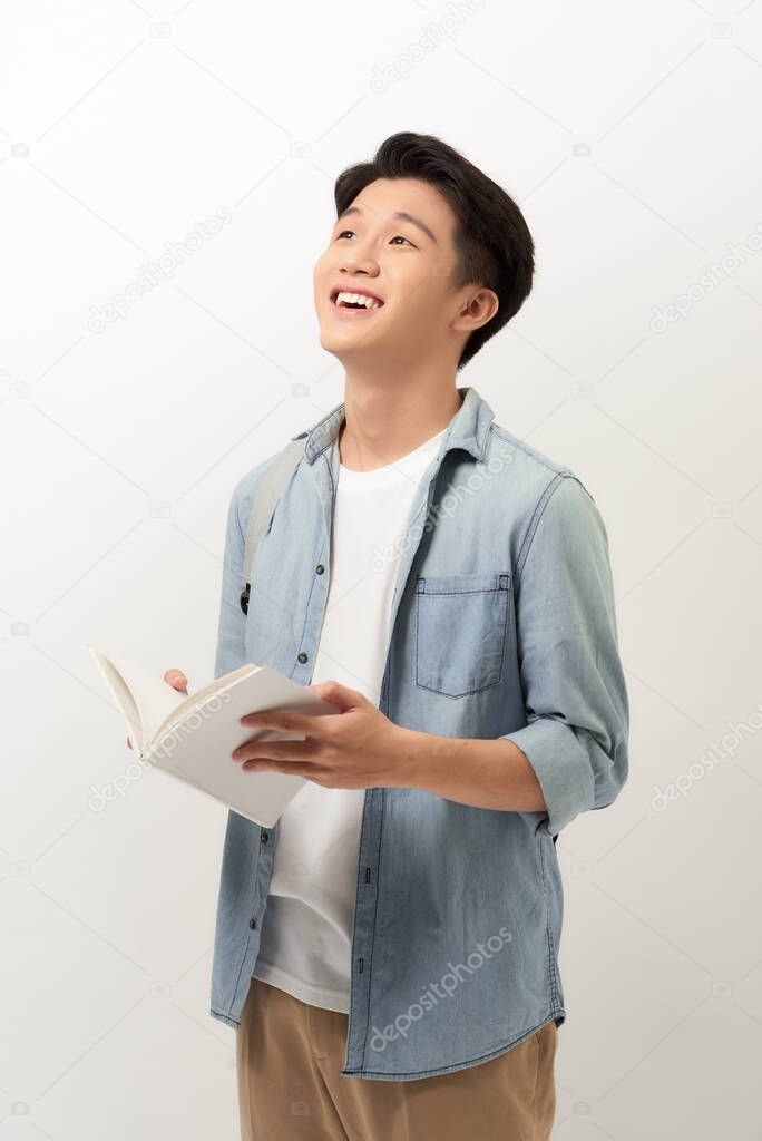 A young Asian man standing in a blue shirt, holding a book and reading, isolated for white background.