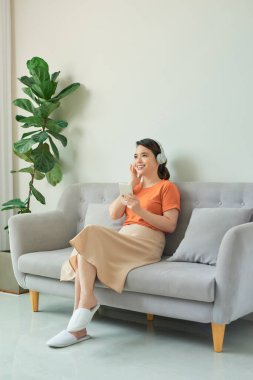 Asyalı çekici kadın kulaklık takıyor, cep telefonu kullanıyor ve koltukta oturuyor.
