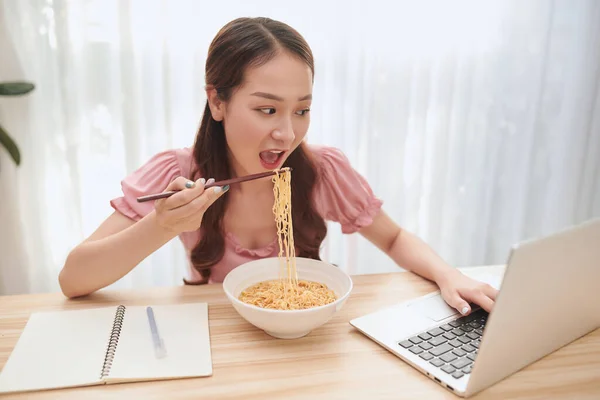 若いアジア人女性食べる麺と作業ノートパソコンで自宅 — ストック写真