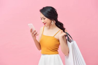 Güzel kız elinde alışveriş torbalarıyla pembe arka planda akıllı bir telefon kullanıyor..