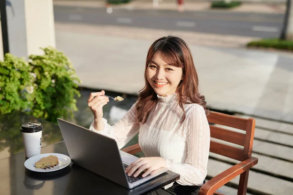 ラップトップを使ってカフェでケーキを食べる陽気な女性 — ストック写真