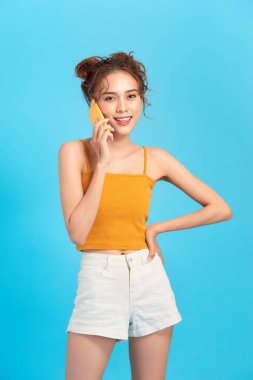 Mutlu gülümseyen genç bir kadın izole edilmiş mavi bir arka planda cep telefonuyla konuşuyor.