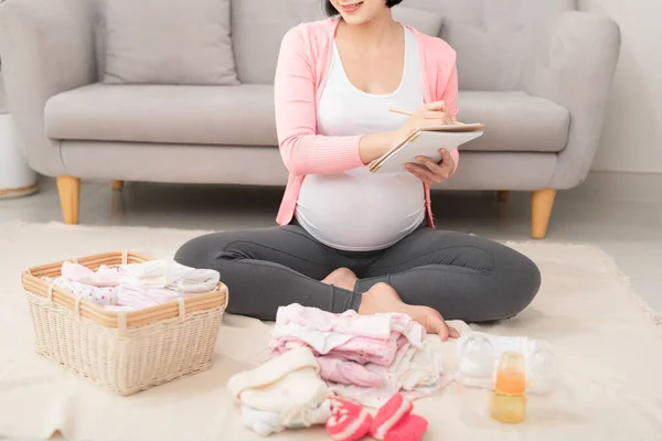 A happy pregnant woman checking a list of things for her unborn baby at home on the floor