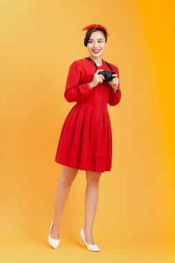 Portakal rengi arka planda dikilirken elinde retro tarzı kamera tutan neşeli, genç Asyalı kadının tüm vücudu..