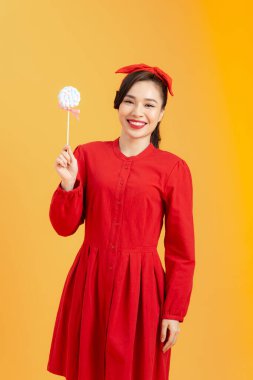 Kırmızı elbiseli neşeli genç Asyalı kadın turuncu arka planda lolipop tutuyor..