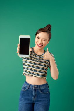 Yeşil arka planda dikilirken boş ekran cep telefonu gösteren sıradan tarzda güzel bir Asyalı kadın..