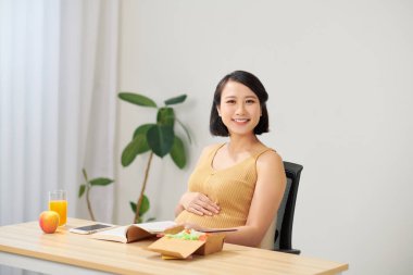 Oturma odasındaki masada oturup dijital tablet kullanan hamile bir kadın kahvaltı yapıp çalışıyor.