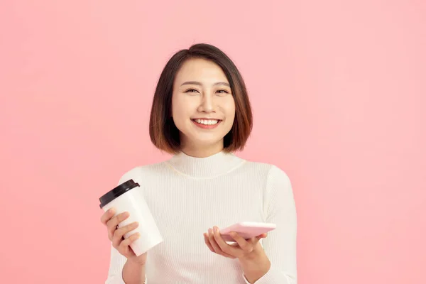 ピンクの背景の上にコーヒーカップとスマートフォンを保持している若いアジアの女性のクローズアップ肖像画 — ストック写真