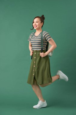 Yeşil arka planda sırt çantasıyla zıplayan ve gülen güzel bir kız öğrenci.