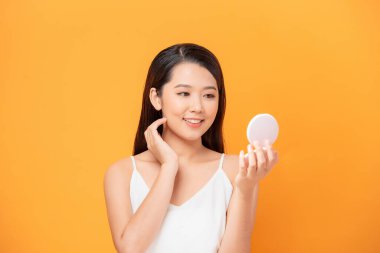 Sarı arka planda izole edilmiş aynaya bakarken yüzünü inceleyen neşeli ve çekici bir kadının güzel portresi.