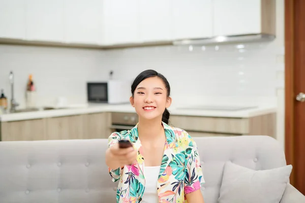 Smiling asian woman on couch changing tv channel at home in the living room