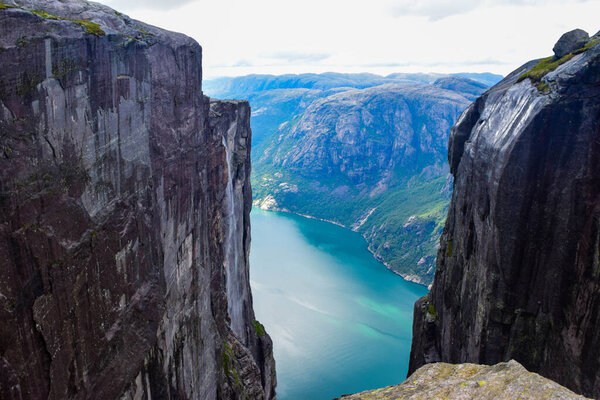 Вид на Люсе-фьорд через трещину между двумя скалами высотой 984 метра, где рядом застрял знаменитый Керагболтен - самый опасный камень в мире. Горный Кьераг, графство Рогаланд, Норвегия
.