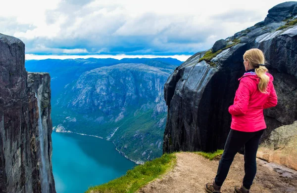 Tourist girl stands on the edge of a cliff 984 meters high above Lysefjorden, where famous Kjeragbolten stuck nearby - the most dangerous stone in the world. Mountain Kjerag, Rogaland county, Norway.