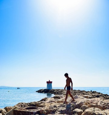 Dalgakıranın taşları üzerinde genç bir adamın silueti, sonunda bir deniz feneri duruyor. Sakin deniz ve mavi gökyüzünün yaz manzarası. Villeneuve-Loubet. Gökyüzü Sahili, Fransız Rivierası.