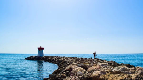 Dalgakıranın taşları üzerinde genç bir adamın silueti, sonunda bir deniz feneri duruyor. Sakin deniz ve mavi gökyüzünün yaz manzarası. Villeneuve-Loubet. Gökyüzü Sahili, Fransız Rivierası.