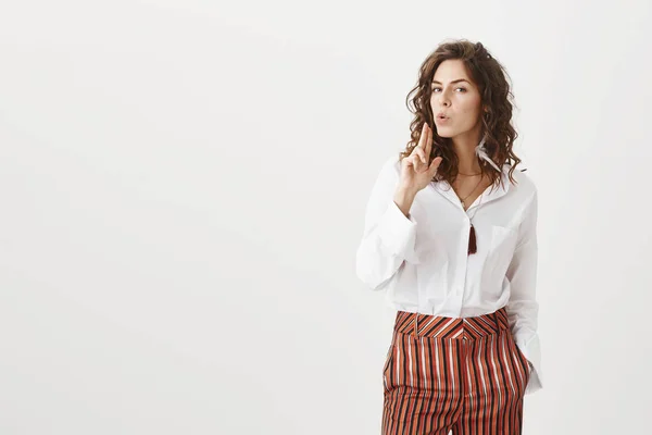 Confident attractive adult woman in stylish striped pants and blouse, showing finger fun gesture near mouth, blowing gunpoint, staring self-assured and serious at camera, being secret agent or spy