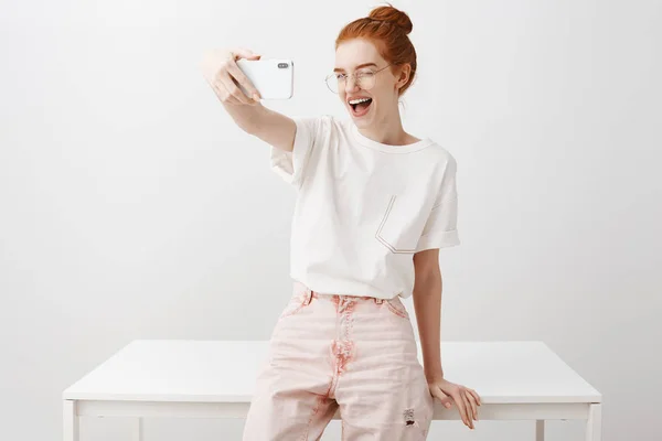 Stylish fashion blogger starting live to talk with followers. Feminine redhead female student in trendy outfit and glasses, leaning on table, taking selfie on smartphone, winking and smiling at screen