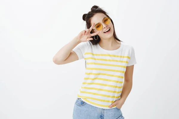 Peace my friends, let us have disco party. Portrait of stylish confident and happy caucasian woman in sunglasses and t-shirt, holding hand in pocket showing victory sign over eye and smiling broadly