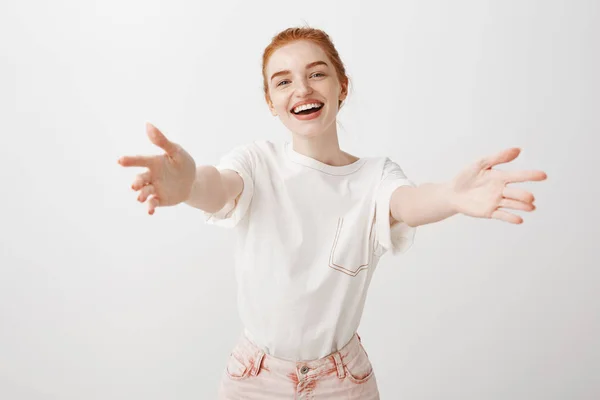 Come to me in warm hugs. Portrait of charming carefree and friendly stylish redhead in pink jeans and t-shirt, pulling hands towards camera to cuddle friend, smiling friendly over gray background