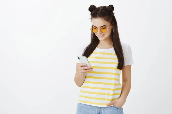 Girl cannot live without internet and social networks. Portrait of stylish carefree woman in sunglasses and t-shirt, holding hand in pocket while scrolling feed in smartphone, smiling joyfully