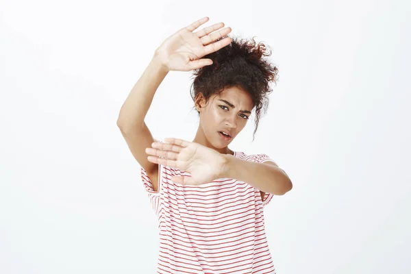 Shut camera off. Portrait of displeased unhappy female student in striped t-shirt, pulling hands towards camera, covering face from paparazzi, standing unhappy and intense over gray background