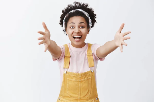 Hey come here and hug me. Indoor shot of charming caring friendly-looking dark-skinned woman in headband and overalls pulling hands towards camera to cuddle best friend, smiling joyfully