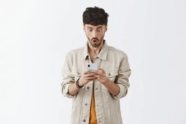 Waist-up shot of stunned amazed and surprised good-looking bearded guy receiving incredible offer via mail reading message on smartphone saying wow popping eyes at device screen from excitement