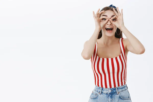 Let us have fun. Funny enthusiastic attractive young woman in striped top and shorts making goggles with fingers over eyes and looking through at camera joyfully smiling broadly from amusement