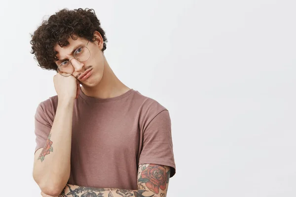 Attractive and stylish young guy with moustache curly hair and tattooed arm leaning on fist pouting and sulking frowning while offended being angry and gloomy friends did not invite to party