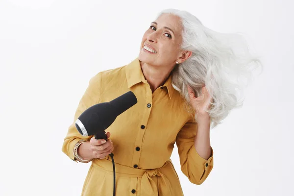 Charming elegant grandmother with grey hair standing in trendy yellow coat bending right, making last preparations to go meet friend drying hair with hairdryer smiling joyfully over gray wall.
