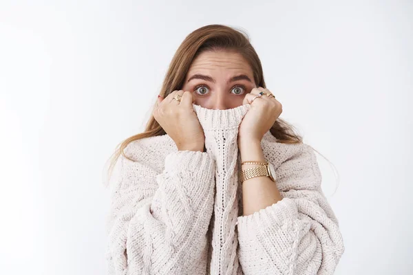 Shocked scared stunned cute caucasian woman hiding face collar sweater pulling cloth nose widen eyes amazed speechless afraid watching horror standing stuned white background, frightened